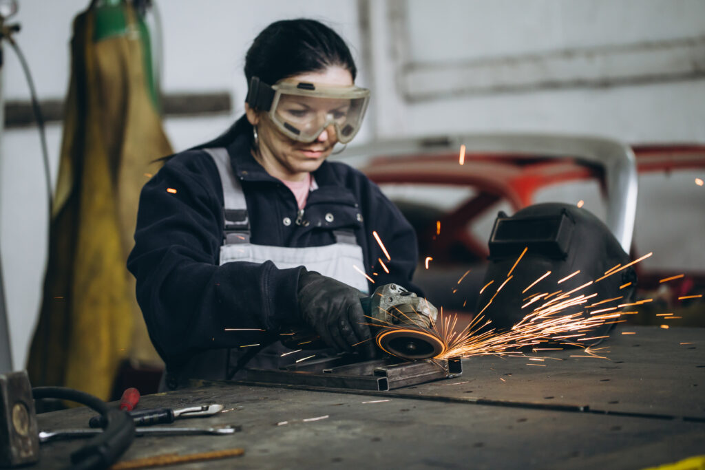 Symbolbild für die Metallbranche: Frau in Arbeitskleidung mit einer Schutzbrille auf, die gerade etwas lötet oder schweißt. Im Hintergrund eine verschwommene Werkstatt-Szenarie.