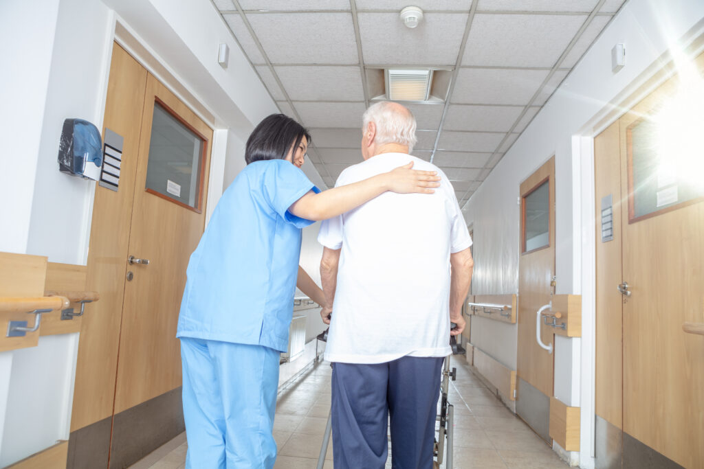 Symbolbild für die Pflegebranche: weibliche Pflegekraft mit grauen Haaren, die einem älteren Mann mit einem Rollator freundlich die Hand auf den Rücken legt. Sie befinden sich im Gang eines Pflegeheims.