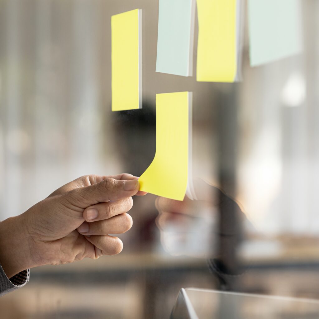 Symbolbild von mehreren leeren Post-It-Zetteln, die an einer Glasscheibe hängen, eine Hand greift nach einem dieser Zettel, im Hintergrund ein verschwommenes Büro-Setting