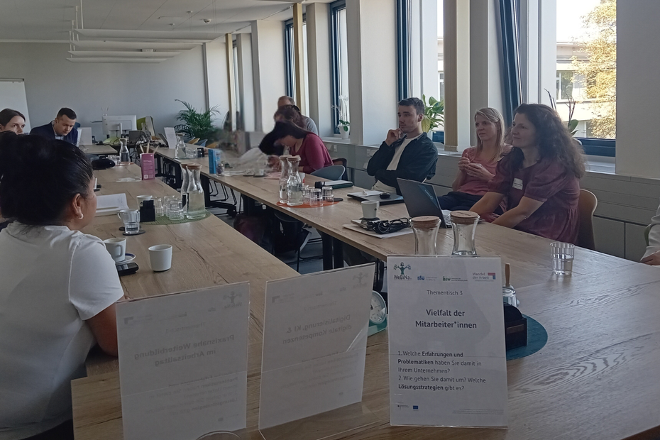 Ein großer Tisch in einem Seminarraum, an dem einige Menschen sitzen, die sich unterhalten. Ein Schild im Vordergrund sagt: "Thementisch 3: Vielfalt der Mitarbeiter*innen". Die Menschen aus den WeBiNa 1.0-Partnerunternehmen sind einander zugewandt, einige beugen sich über den Tisch und schreiben etwas auf.